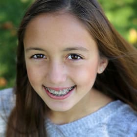 Young girl with braces smiling at camera outdoors with green foliage background.