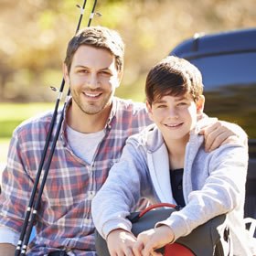 Father and son smiling while holding fishing rods outdoors near a vehicle.