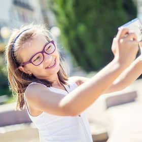 Young girl wearing pink glasses smiling while taking selfie outdoors in sunny garden setting.