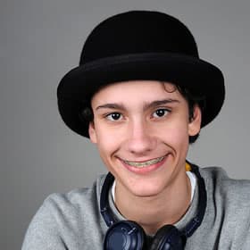 Young man wearing black bowler hat and gray shirt, smiling at camera against gray background.