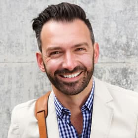 Professional headshot of smiling man in white blazer against textured gray wall background