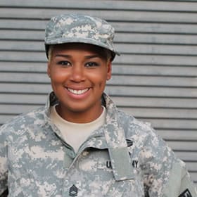 Smiling soldier in digital camouflage uniform and cap against corrugated metal background.
